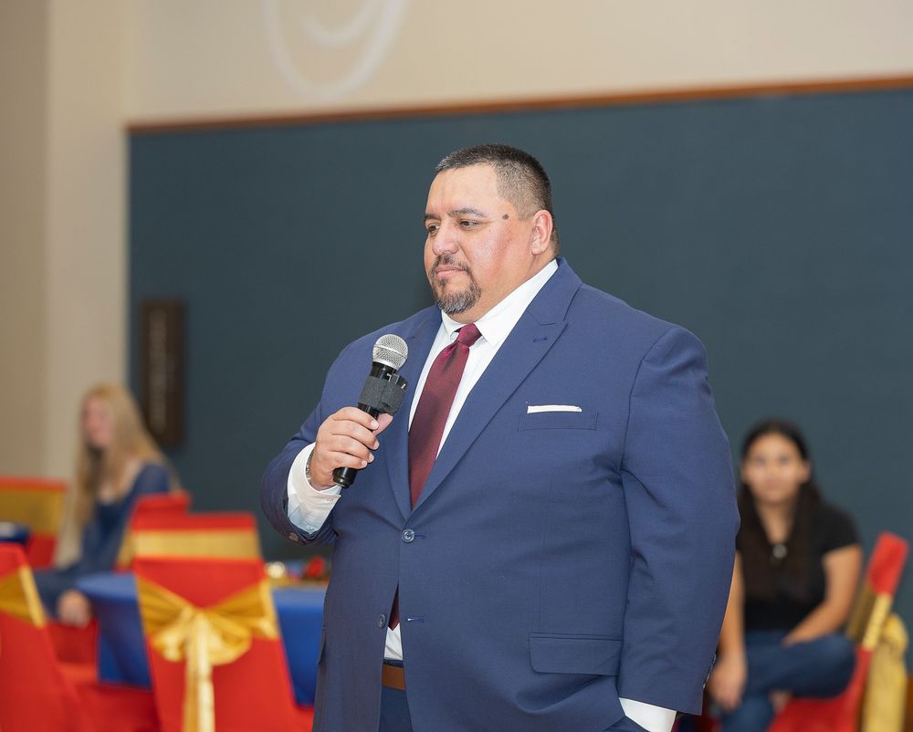Speaker in a navy blue suit holds a microphone while presenting at an indoor event with red and gold decorations.
