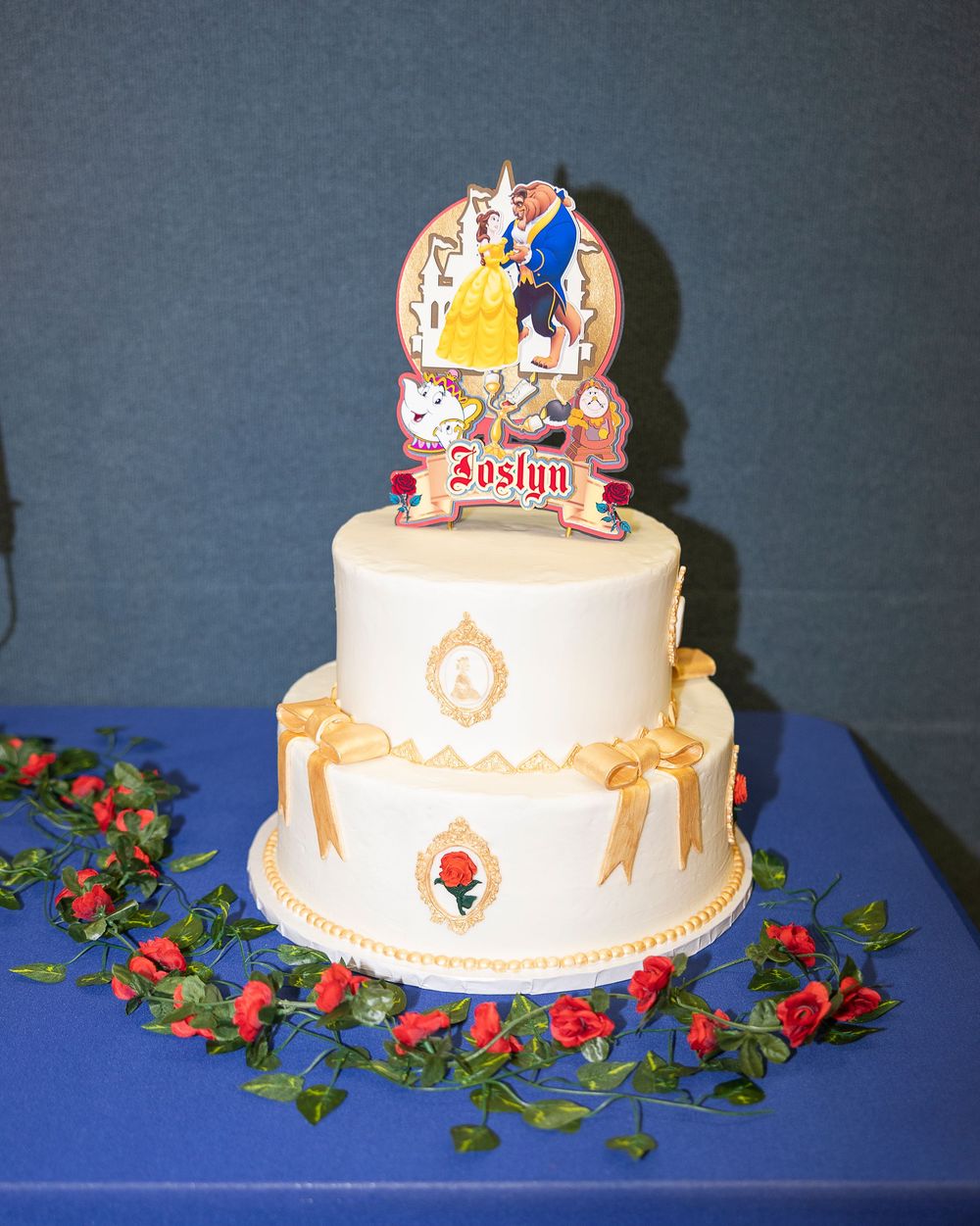 A white two-tiered wedding cake decorated with gold accents and topped with a decorative crown on a blue tablecloth.