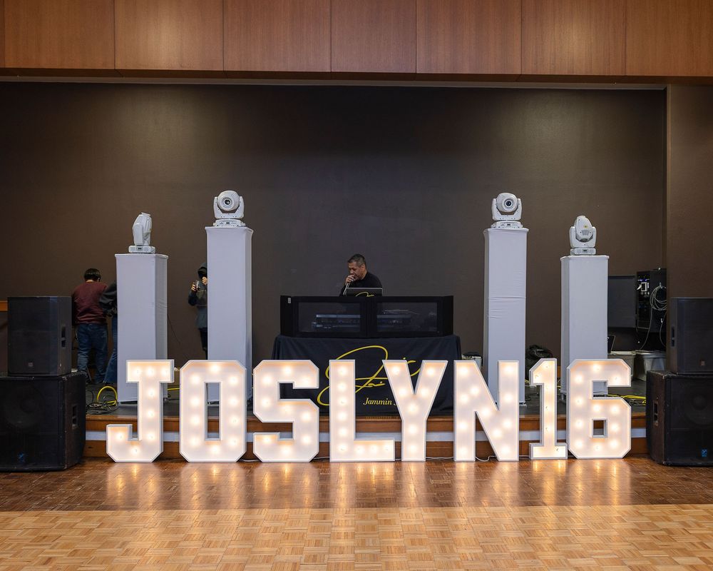 Large illuminated letters spelling 'JOSLYN16' displayed on a wooden floor with white pedestals and DJ setup in background.