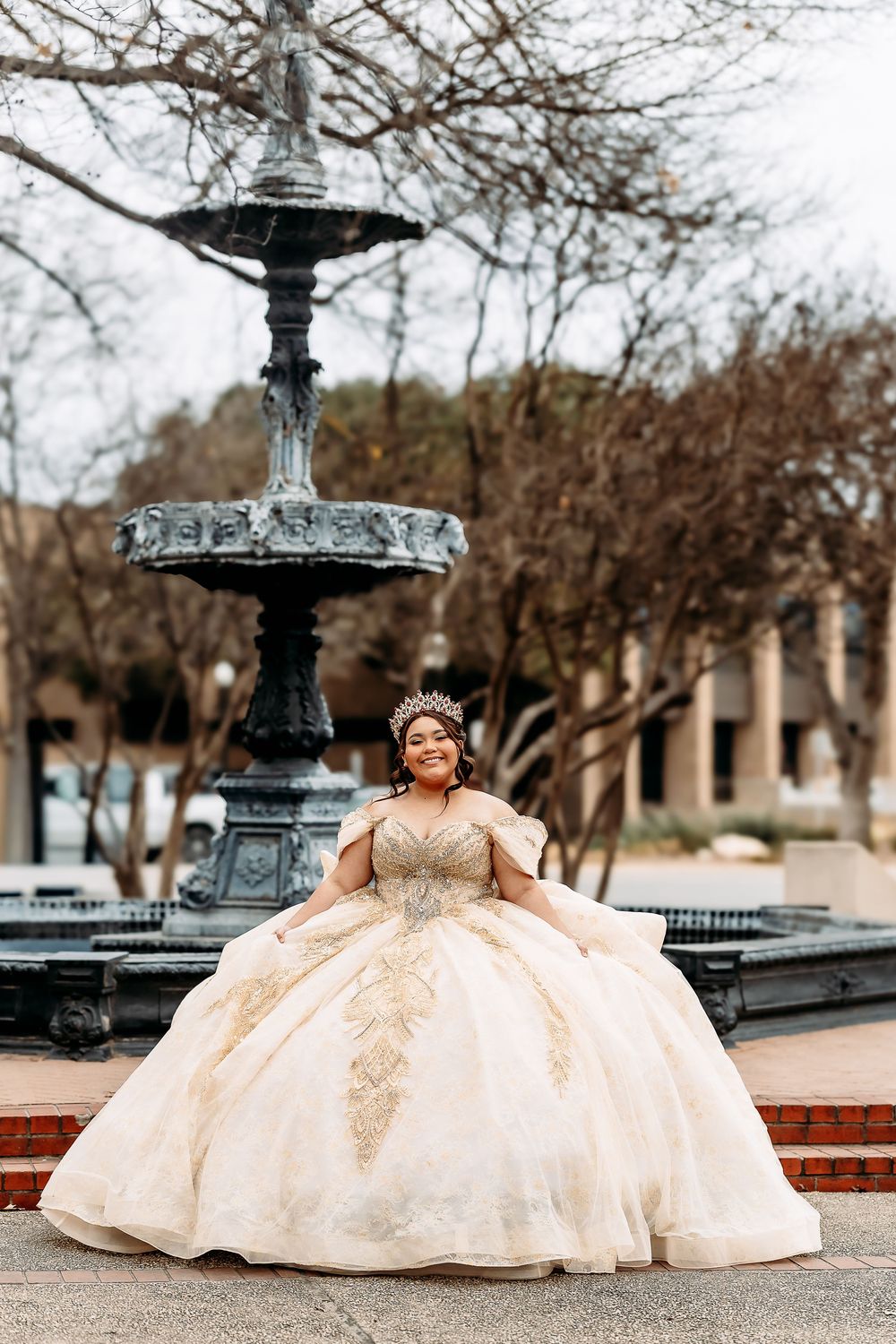 An ornate cream-colored ballgown with gold embellishments photographed in front of a decorative fountain in a park setting.