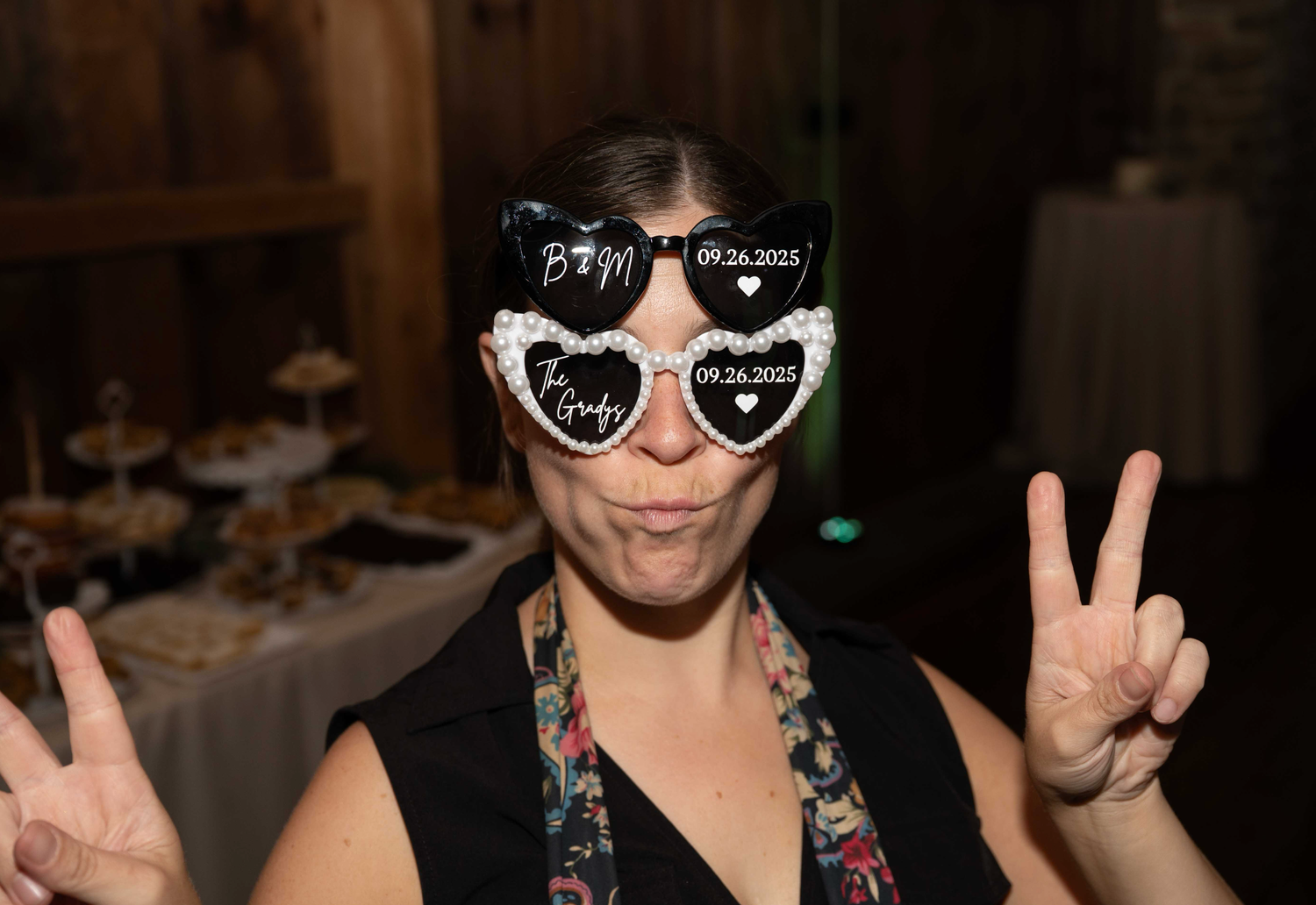 Person wearing heart-shaped sunglasses and floral print making peace signs in a dimly lit setting.