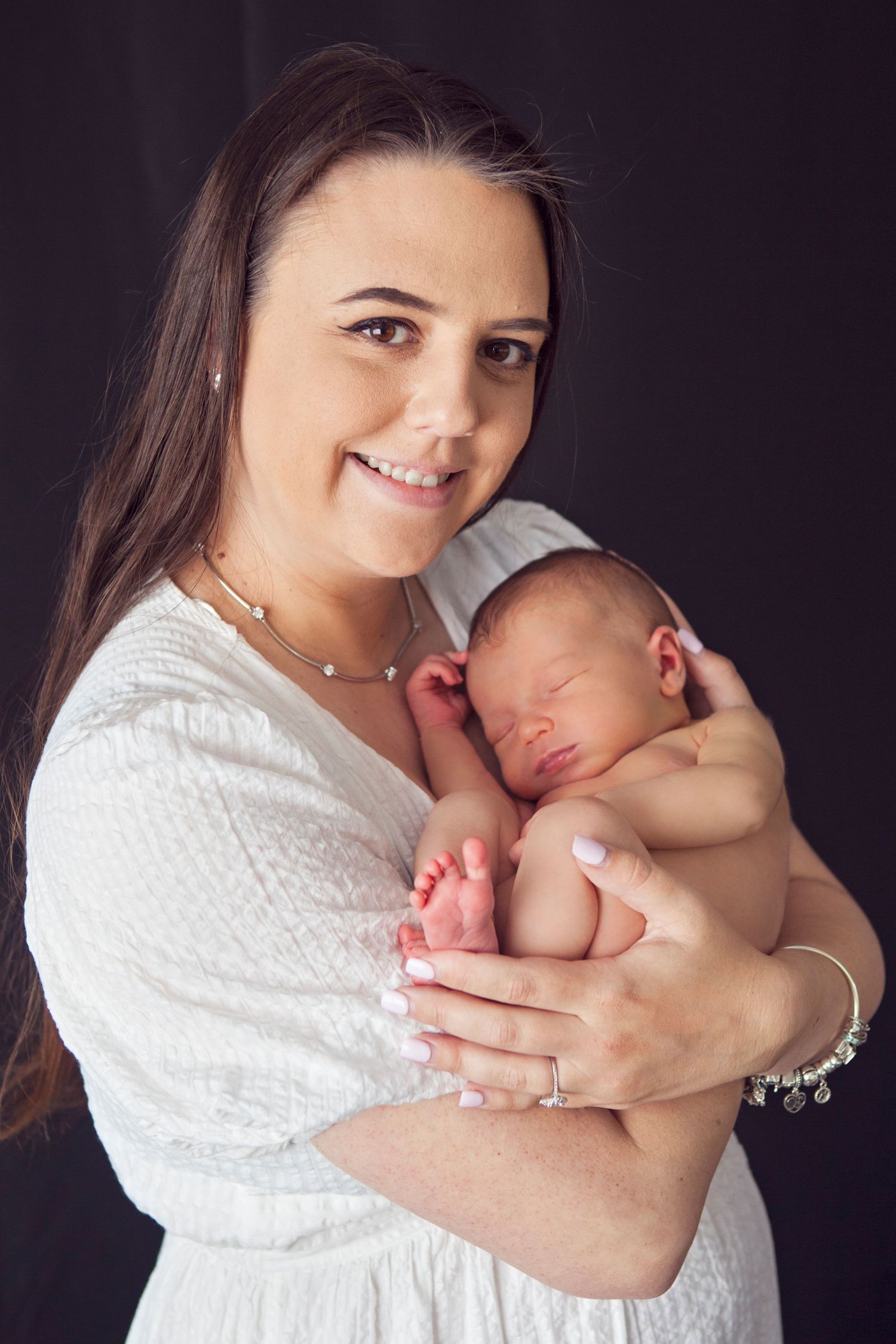 Smiling brunette mother in white dress holding sleeping newborn baby against dark background.