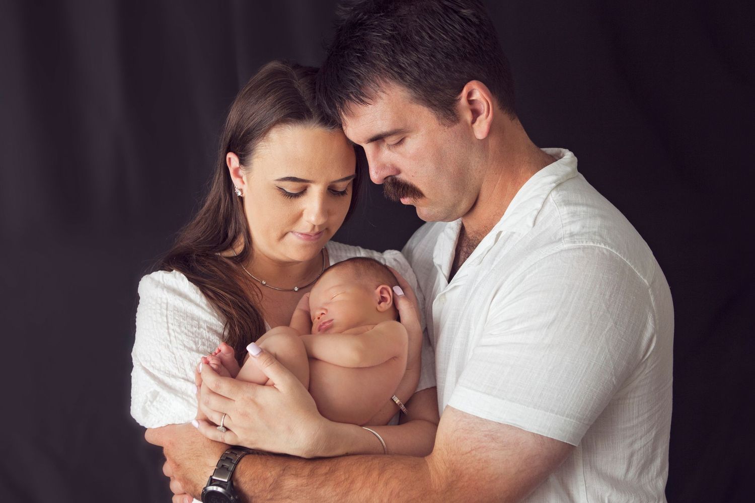 Parents tenderly cradling their sleeping newborn baby together against a dark studio background.