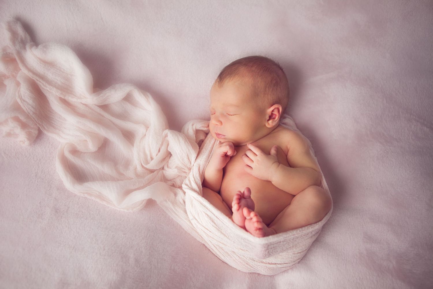 Peaceful newborn baby sleeping wrapped in soft white cloth on pink blanket in professional studio photography.