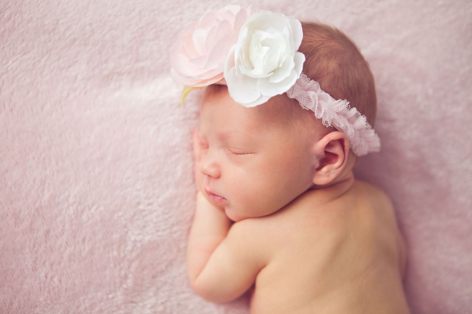 Sleeping newborn baby girl wearing a delicate pink and white floral headband on a soft pink blanket.