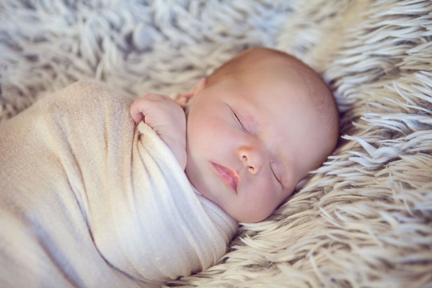 A peaceful sleeping baby swaddled in white lies on soft white fur bedding.
