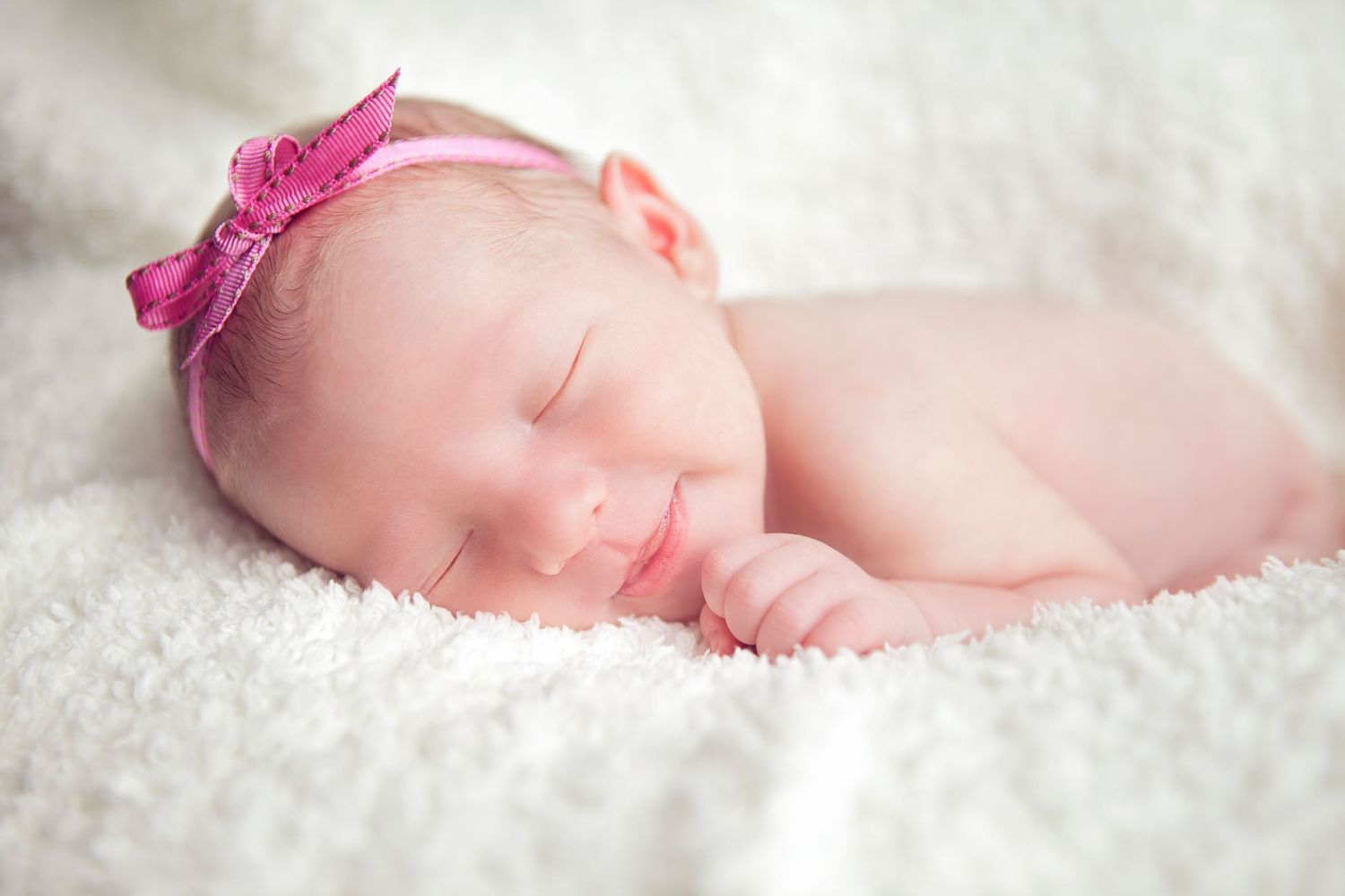 A newborn baby sleeping peacefully on soft white blankets with a pink bow headband.