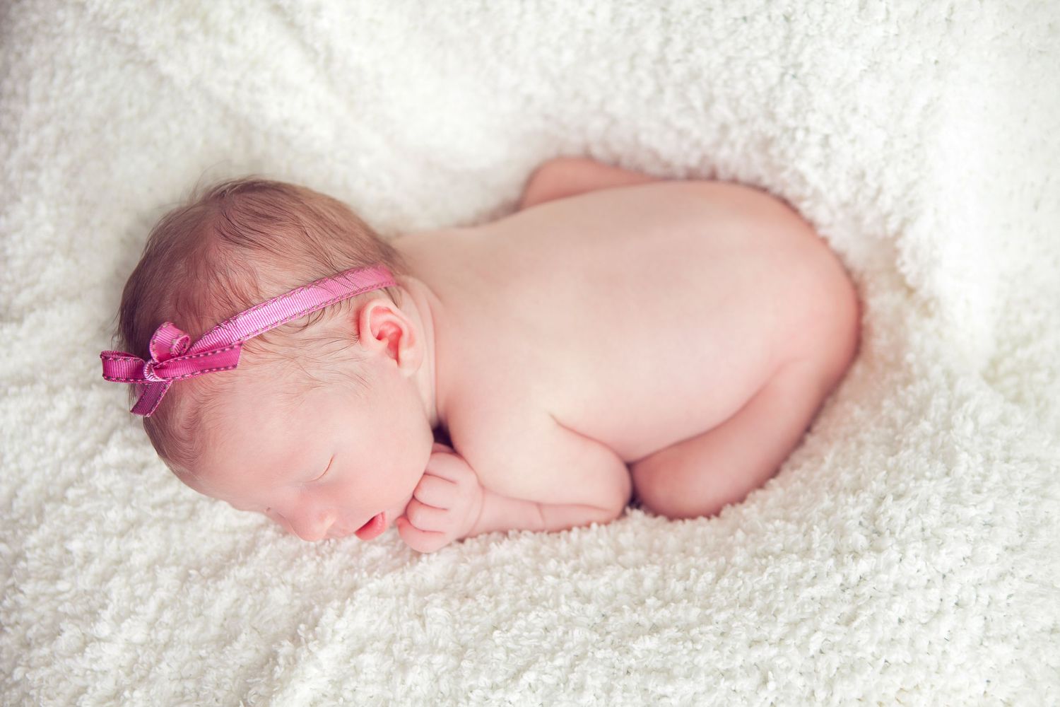 Sleeping newborn baby wearing pink headband curled up on soft white blanket.