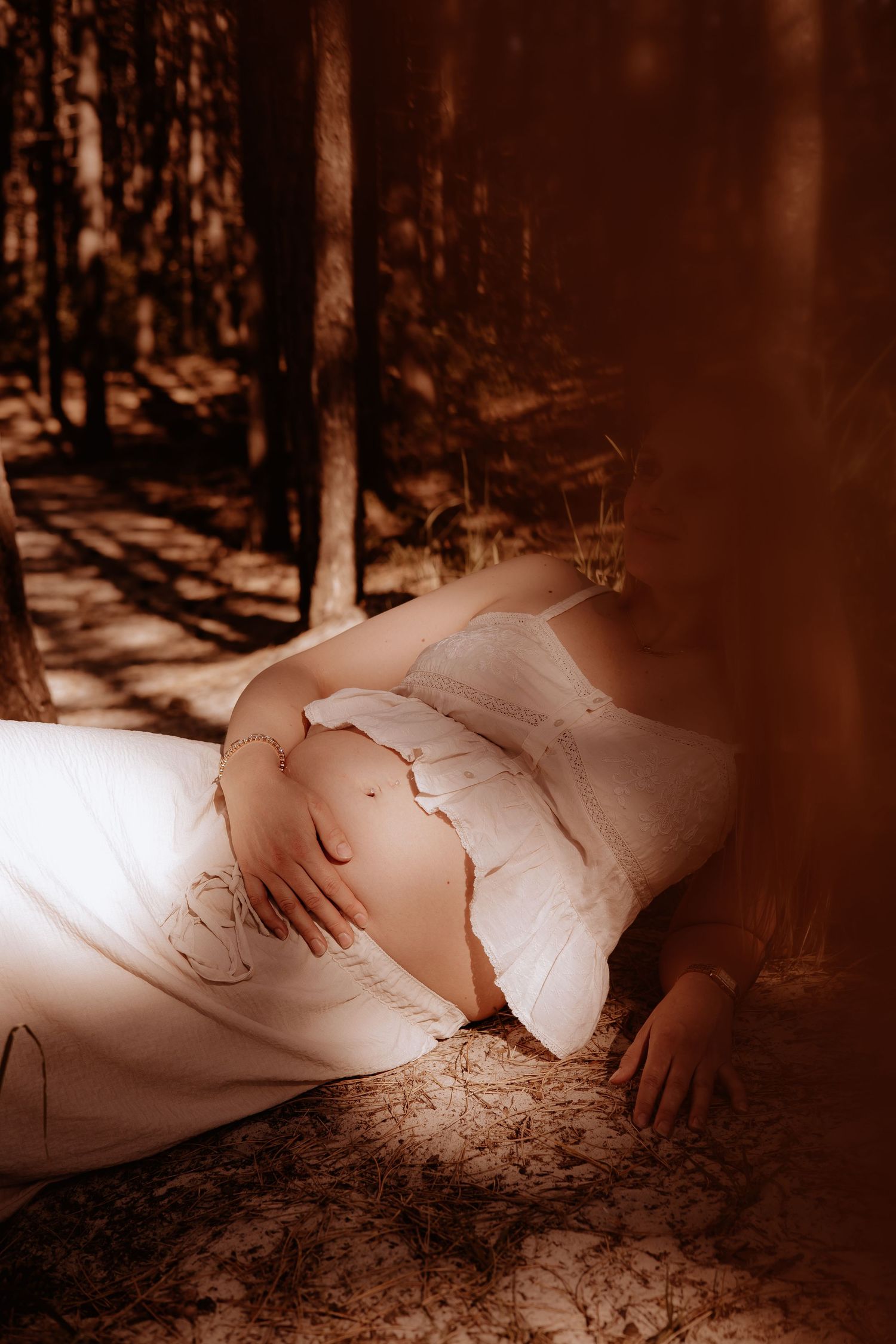 Pregnant woman in white dress lying against a tree in sunlit forest, cradling her bare baby bump.
