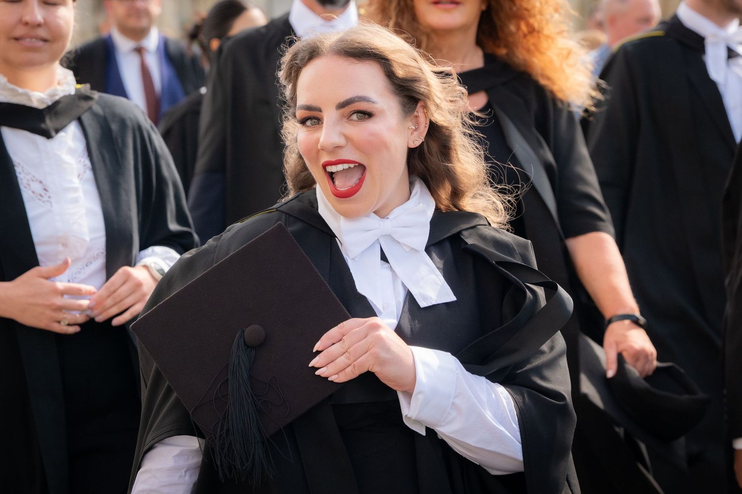 Excited graduate in black academic gown and cap celebrating at graduation ceremony outdoors.
