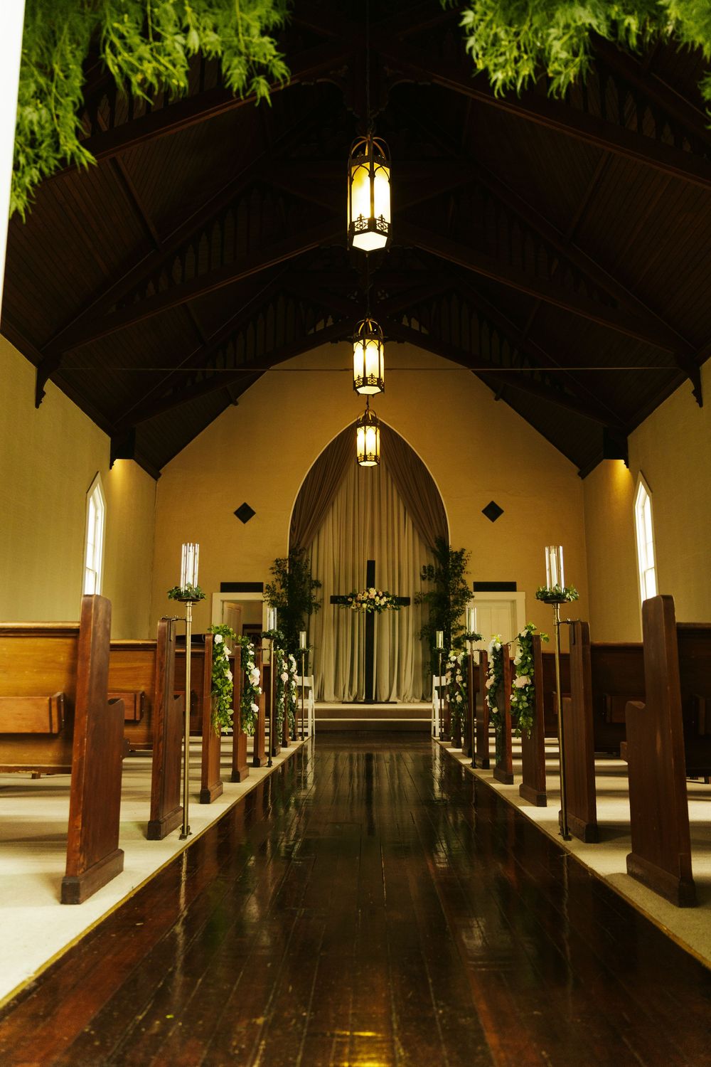 Elegant chapel interior with wooden pews, hanging lanterns, floral decorations, and a draped altar at the end of a dark aisle.