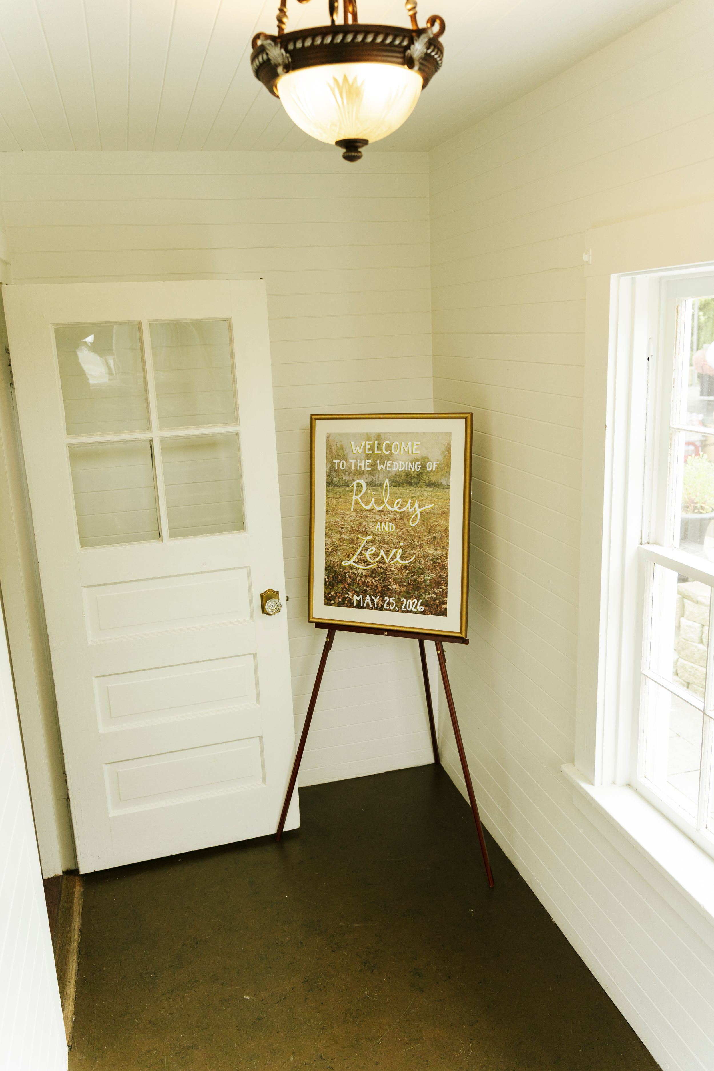 Wedding welcome sign on easel in bright white entryway with glass panel door and pendant light.