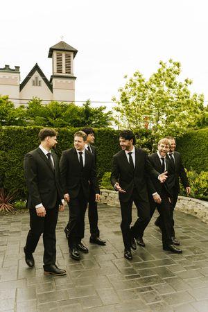 Six groomsmen in black suits and ties walk together laughing outside a church on a wedding day.