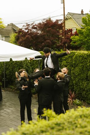 Groomsmen in black suits lifting groom outdoors at wedding reception in garden setting.