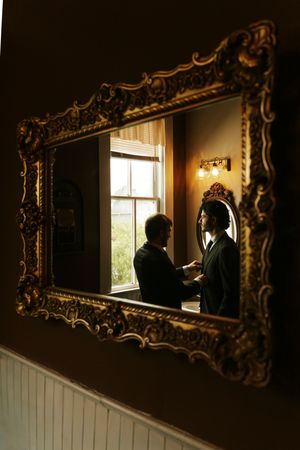 Ornate gold mirror reflecting two men in silhouette near a bright window in a warmly lit room.