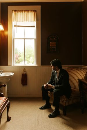 A groom in a dark suit sits pensively on a striped chair by a window in a vintage-styled room.