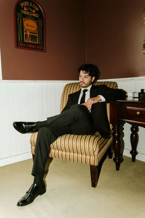 Man in black suit sitting relaxed in striped armchair near wooden table, with tobacconist sign on wall.