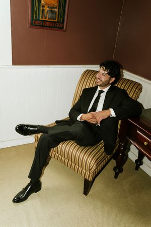 A man in a black suit and tie sits relaxed in a striped armchair, legs crossed, in a room with dark red walls.