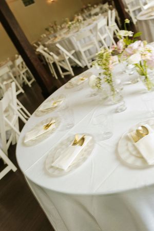 Elegant wedding reception tables with white linens, gold cutlery, marble chargers, and floral centerpieces in a softly lit venue.
