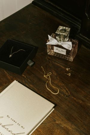 Elegant bridal flat lay with Miss Dior perfume, gold jewelry, necklace in box, and vow book on dark wood.