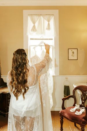 Bride in lace robe holding wedding dress on hanger near sunlit window in vintage room with wooden chair.