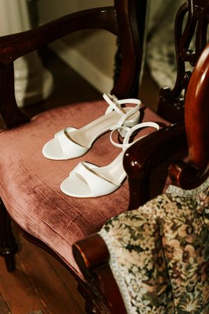 White bridal sandals with ankle straps displayed on a vintage rose velvet chair with dark wood arms.
