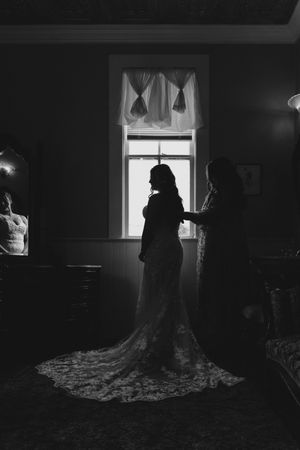 Black and white silhouette of bride in lace gown with train standing by window while bridesmaid helps her dress.
