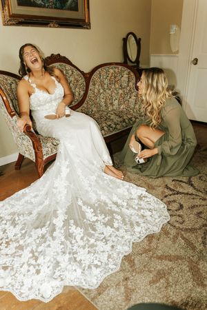 A bride in a lace wedding gown laughs joyfully as her bridesmaid helps her put on shoes before the ceremony.