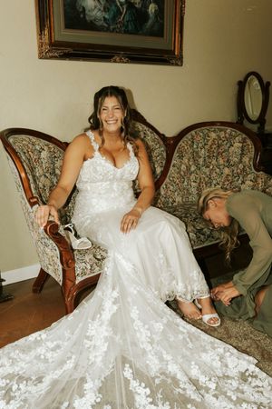 Bride in lace wedding gown sits on ornate floral sofa smiling while bridesmaid helps put on white heels.