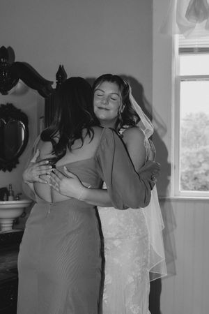 Black and white photo of a bride in a lace gown and veil sharing a tender embrace with a bridesmaid in a satin dress.