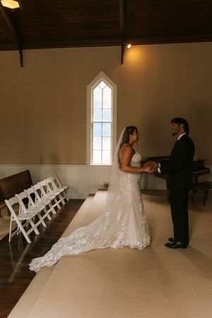 Bride and groom holding hands in a white chapel with arched windows and white folding chairs during wedding ceremony.