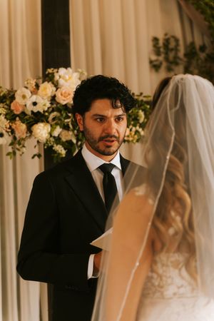 Groom in black suit gazes lovingly at bride in white veil during wedding ceremony with floral backdrop.