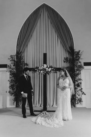 Black and white photo of bride and groom exchanging vows before a floral cross altar with draped arch backdrop.