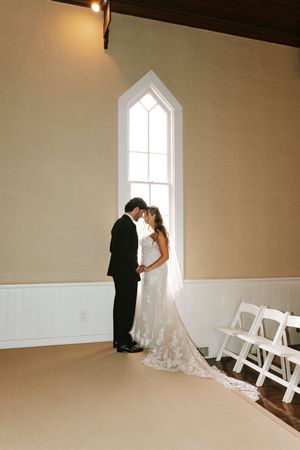 Bride and groom share an intimate moment in a sunlit chapel, with a lace wedding gown train trailing on the floor.