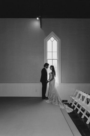 Black and white photo of a bride and groom holding hands by a chapel window, their silhouettes glowing in soft natural light.