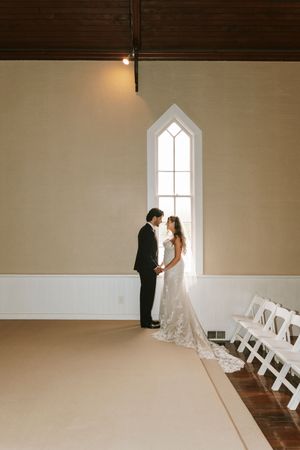 Bride and groom holding hands near tall arched window in elegant white chapel during wedding ceremony.