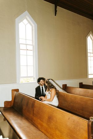 Bride and groom share an intimate moment seated together in wooden church pews near sunlit chapel windows.
