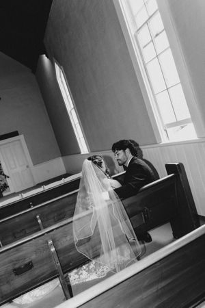Black and white photo of bride and groom sitting together in church pew, bride wearing veil.