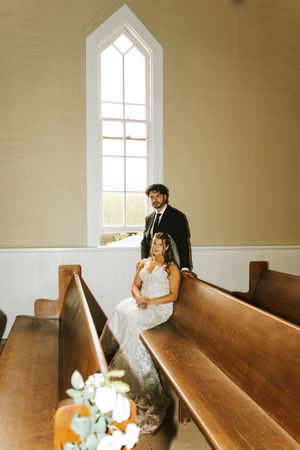Bride in lace gown sits on wooden church pew beside groom in black suit near sunlit chapel window.