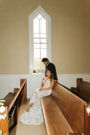 Bride and groom posing in church pew beside tall arched window on wedding day.