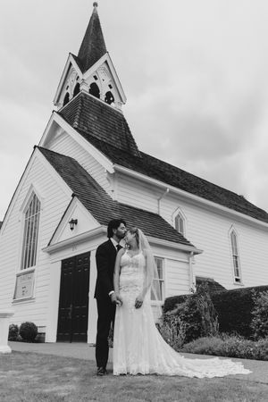 Black and white photo of a bride and groom kissing in front of a white chapel with a steeple on their wedding day.