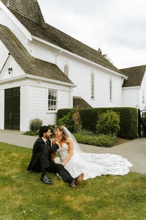 Bride and groom sitting together on grass outside a white chapel on their wedding day.