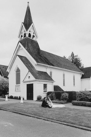 Black and white photo of bride and groom seated outside a white church with a tall steeple.