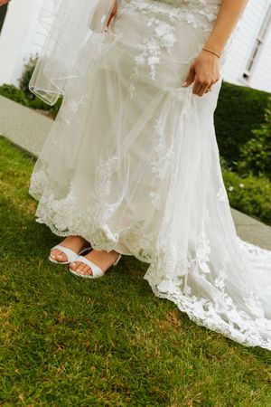 Close-up of bride's lace wedding gown train and white heeled sandals on green grass.