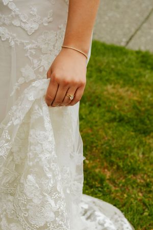 Bride in lace wedding gown holding dress hem, wearing gold diamond engagement ring and delicate bangle bracelet outdoors.