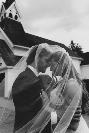 Black and white photo of bride and groom sharing intimate moment under wedding veil outside white chapel.