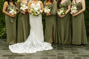 Bride in lace gown with bridesmaids in olive green dresses holding white floral bouquets on stone patio.