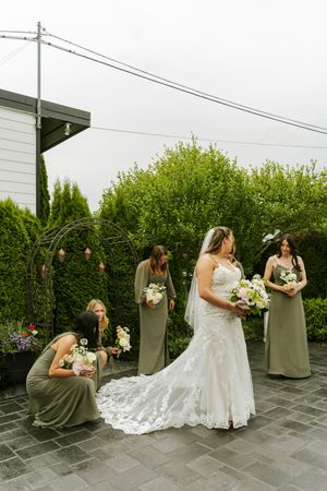 Bride in lace wedding gown with veil holds bouquet while bridesmaids in sage green dresses arrange her train outdoors.