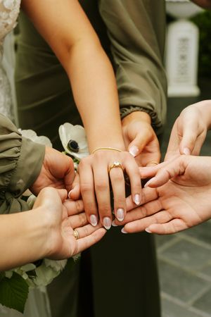 Multiple hands stacked together showcasing an engagement ring with gold band and diamond, with French manicured nails at a wedding.