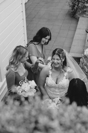 Black and white photo of bride in lace gown laughing with bridesmaids adjusting her veil and jewelry before wedding.